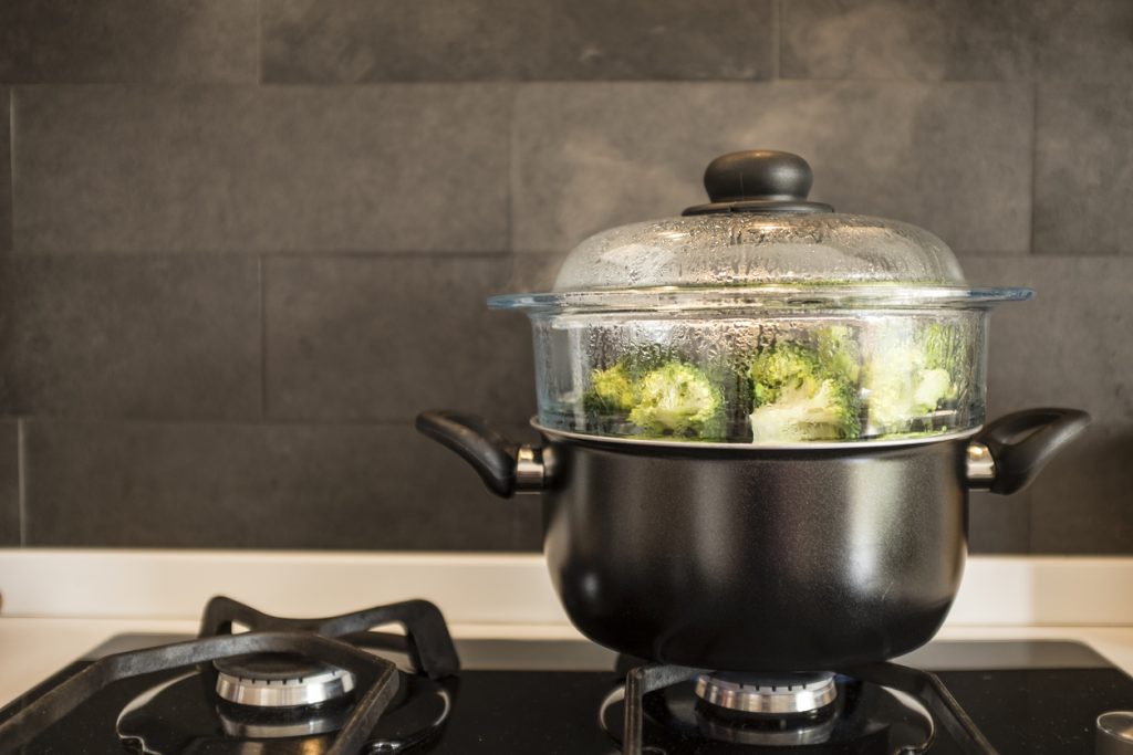 No fogão desligado uma panela de metal, e acima dela uma vasilha de vidro cozinhando o brócolis a vapor.